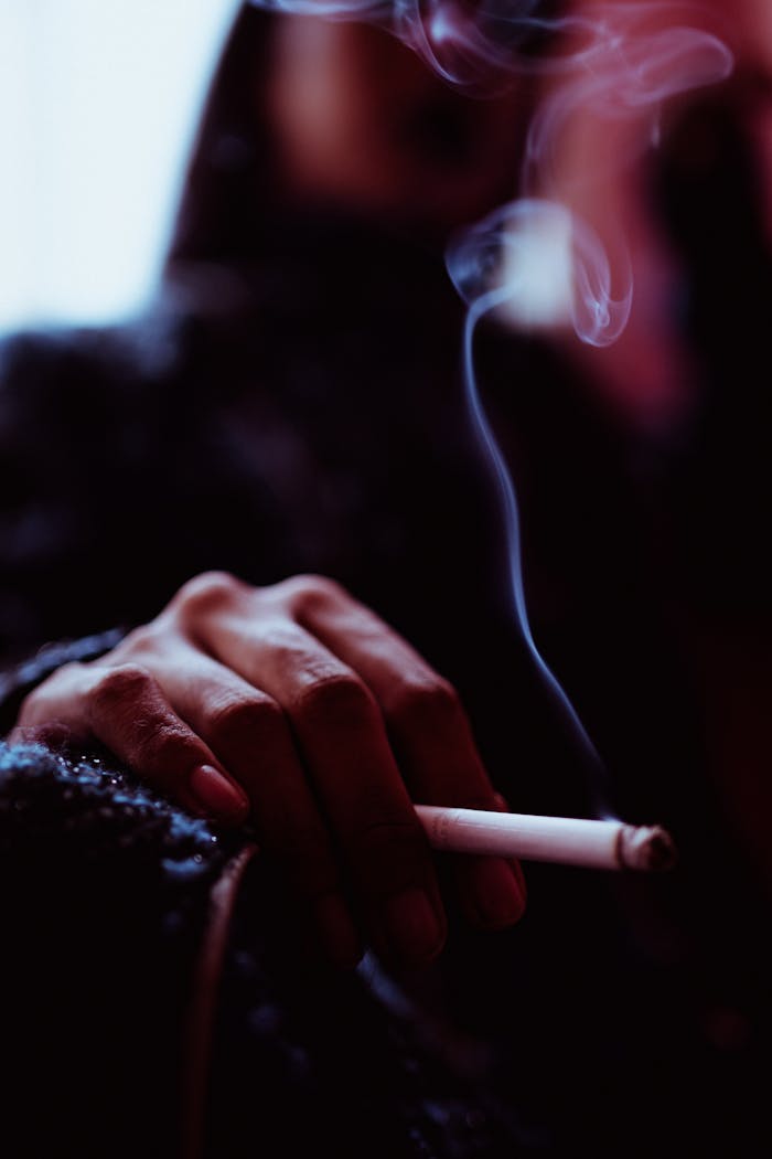 Anonymous blurred female smoker sitting in room near window with smoking cigarette while inhaling fume and relaxing at low light